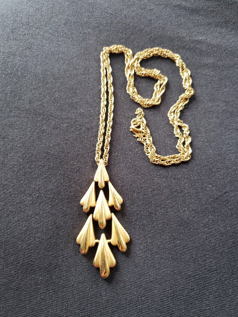 Pu&ograve; includere: Una collana dorata con un pendente a cascata a forma di foglie. La catena &egrave; a corda intrecciata e il pendente presenta molteplici forme di foglie stilizzate a strati. La collana &egrave; esposta su uno sfondo di tessuto grigio scuro.