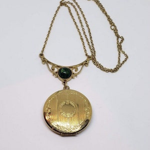 May include: A gold locket necklace with a green gemstone and a detailed filigree design. The locket is round and has a textured surface.