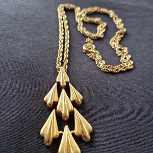 Pu&ograve; includere: Collana color oro con un ciondolo a cascata di elementi stilizzati a forma di foglia. La catena &egrave; un delicato design a corda e il ciondolo ha una superficie strutturata. La collana &egrave; esposta su uno sfondo grigio scuro.