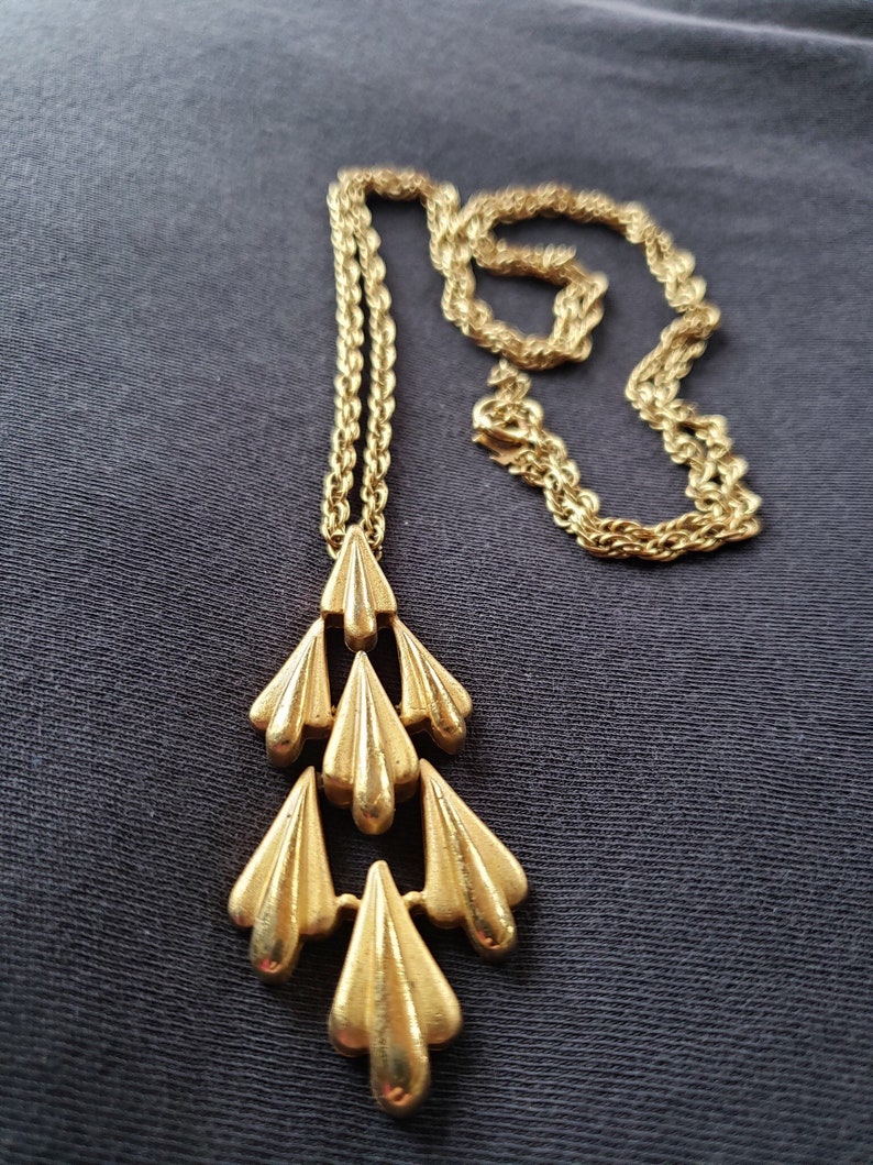 Pu&ograve; includere: Collana color oro con un ciondolo a cascata di elementi stilizzati a forma di foglia. La catena &egrave; un delicato design a corda e il ciondolo ha una superficie strutturata. La collana &egrave; esposta su uno sfondo grigio scuro.
