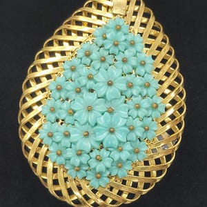 May include: A gold-toned pendant with a woven basket design, shaped like a teardrop. The pendant features a cluster of light blue flower accents with gold-colored centers. The pendant has a loop at the top for hanging.