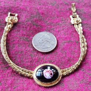 May include: Gold-toned bracelet with a black oval centerpiece featuring a painted rose design. The bracelet has a woven chain design and a clasp closure. A U.S. quarter is next to the bracelet for size comparison.