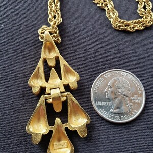 Pu&ograve; includere: Collana Trifari color oro con pendente geometrico. Il pendente ha un design a strati con spazi aperti e una finitura strutturata. La collana &egrave; esposta su sfondo scuro accanto a una moneta come riferimento delle dimensioni.