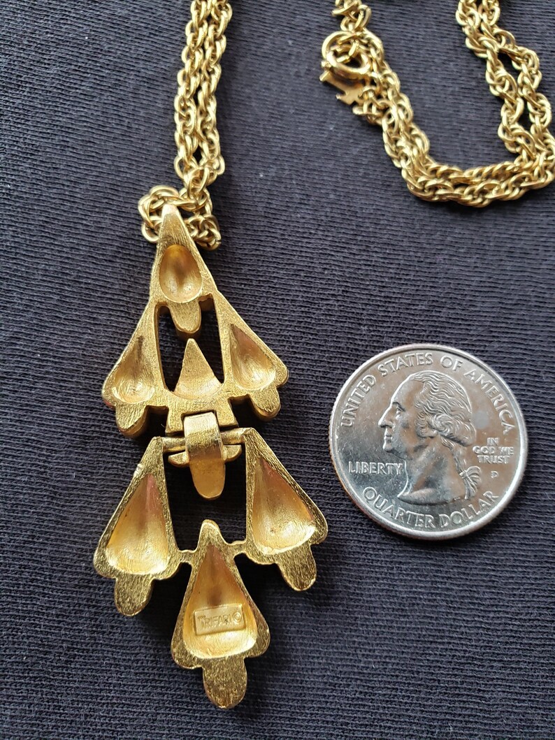 Pu&ograve; includere: Collana Trifari color oro con pendente geometrico. Il pendente ha un design a strati con spazi aperti e una finitura strutturata. La collana &egrave; esposta su sfondo scuro accanto a una moneta come riferimento delle dimensioni.