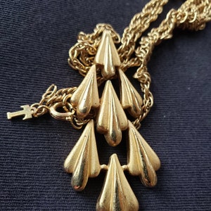 Pu&ograve; includere: Collana color oro con un ciondolo con sei elementi stilizzati a forma di goccia. Il ciondolo &egrave; sospeso a una delicata catena. La collana &egrave; esposta su uno sfondo scuro.