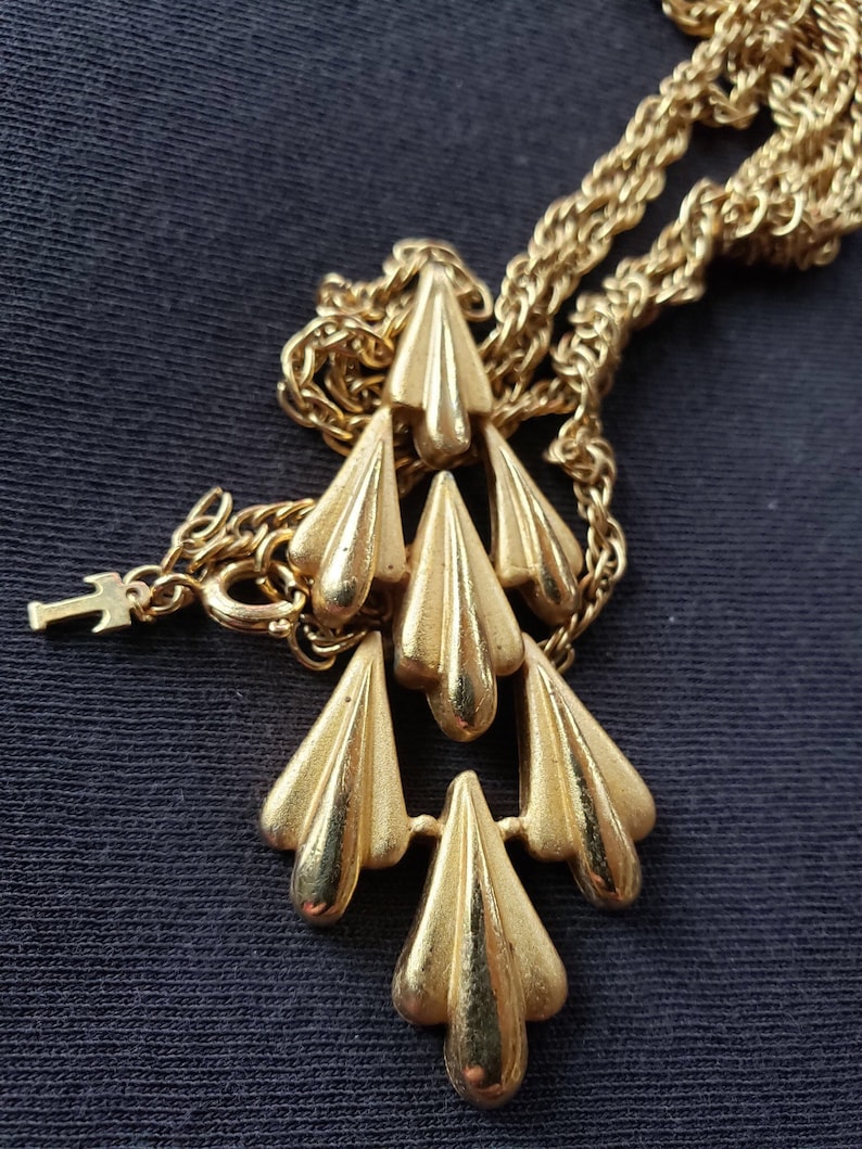Pu&ograve; includere: Collana color oro con un ciondolo con sei elementi stilizzati a forma di goccia. Il ciondolo &egrave; sospeso a una delicata catena. La collana &egrave; esposta su uno sfondo scuro.