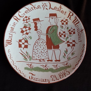 May include: A decorative ceramic plate with a hand-painted design. The plate features a couple in period clothing, surrounded by floral and bird motifs. The names "Marjorie A. Gehrke" and "Lester R. Montout" are inscribed, along with the date "January 21, 1943".
