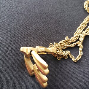 Pu&ograve; includere: Una collana color oro con un ciondolo con un design stilizzato di tre forme a goccia. La catena &egrave; in stile corda ritorta delicata. La collana &egrave; esposta su uno sfondo scuro.