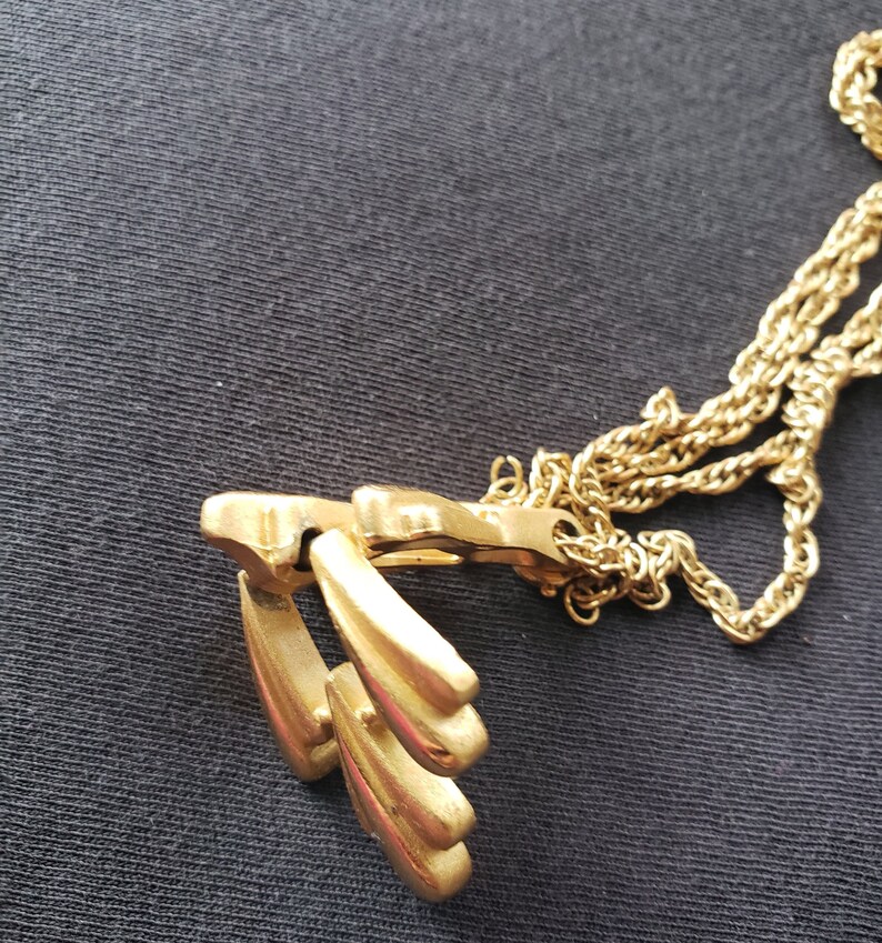 Pu&ograve; includere: Una collana color oro con un ciondolo con un design stilizzato di tre forme a goccia. La catena &egrave; in stile corda ritorta delicata. La collana &egrave; esposta su uno sfondo scuro.