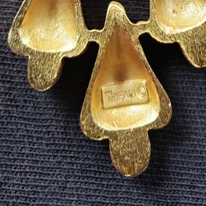 Pu&ograve; includere: Primo piano di una spilla Trifari dorata. La spilla presenta un design floreale stilizzato con tre elementi a forma di petalo. Il nome del marchio Trifari &egrave; visibile sulla spilla.