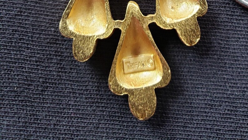 Pu&ograve; includere: Primo piano di una spilla Trifari dorata. La spilla presenta un design floreale stilizzato con tre elementi a forma di petalo. Il nome del marchio Trifari &egrave; visibile sulla spilla.