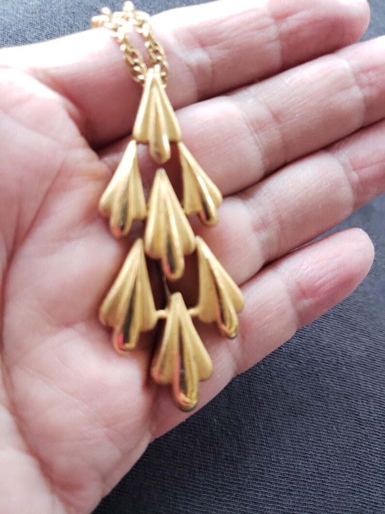 Pu&ograve; includere: Una collana con pendente dorato con un design a cascata di elementi stilizzati a forma di ventaglio. La collana ha una catena delicata e una finitura lucida, creando un'estetica vintage. Il design del pendente &egrave; simmetrico e accattivante.