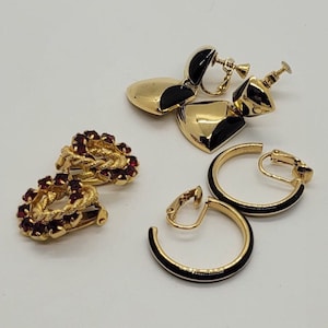 May include: A collection of vintage clip-on earrings in gold tones. Includes heart-shaped earrings with black enamel accents, hoop earrings with black enamel, and earrings with red gemstones. The earrings are displayed on a white background.