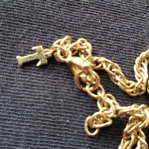 Pu&ograve; includere: Primo piano di una collana color oro con un piccolo ciondolo a forma di croce. La catena ha un design intrecciato e una chiusura a moschettone. La collana &egrave; su uno sfondo scuro.