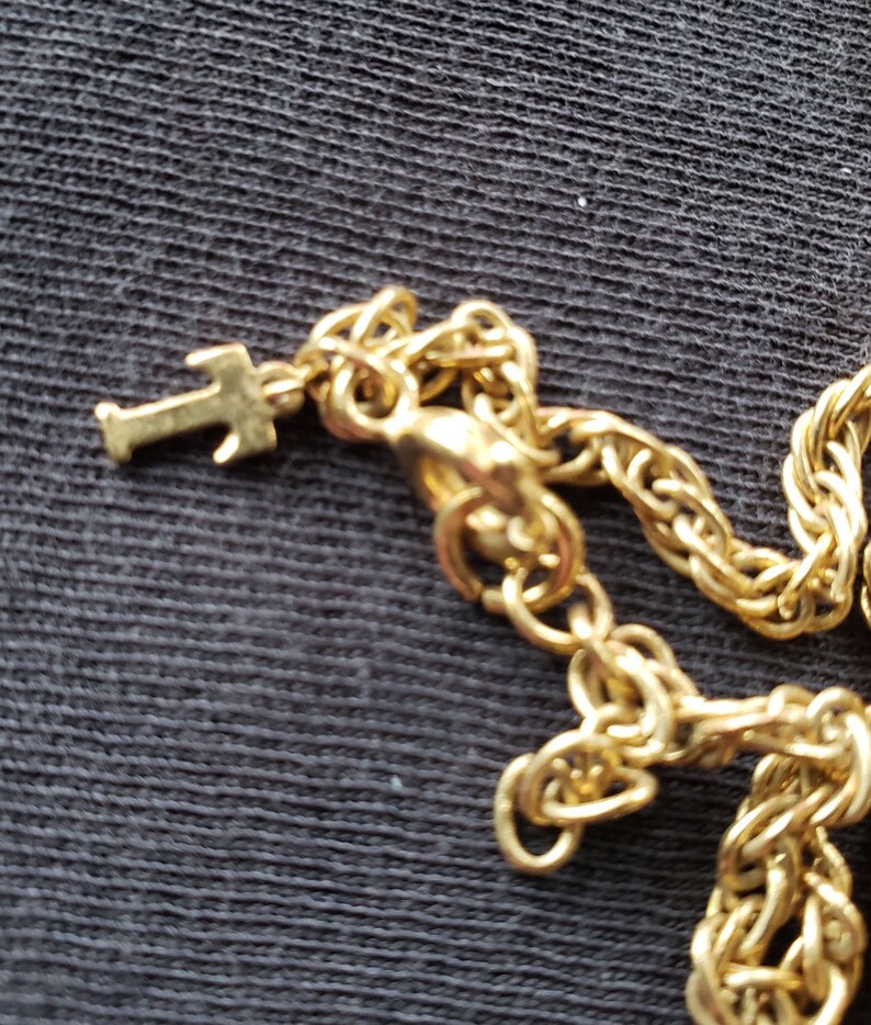 Pu&ograve; includere: Primo piano di una collana color oro con un piccolo ciondolo a forma di croce. La catena ha un design intrecciato e una chiusura a moschettone. La collana &egrave; su uno sfondo scuro.