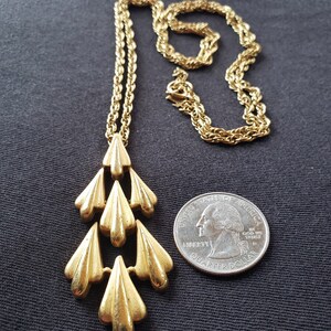 Pu&ograve; includere: Una collana dorata con un pendente a cascata di elementi stilizzati a forma di foglia. La catena &egrave; in stile corda classico. Un quarto di dollaro americano &egrave; incluso per riferimento alle dimensioni. La collana &egrave; esposta su sfondo scuro.