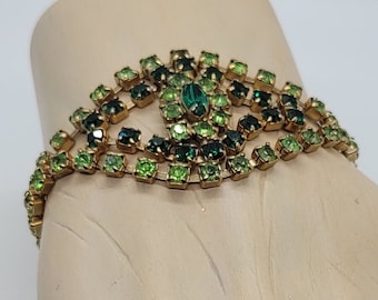 May include: A vintage bracelet with a gold-tone metal frame. The bracelet features a series of green and dark green square-cut rhinestones. A large oval green rhinestone is at the center. The bracelet is on a mannequin hand.