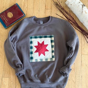 May include: A charcoal gray crewneck sweatshirt featuring a square fabric patch on the chest. The patch has a white quilted center with a red eight-pointed star and a dark green and white gingham border, attached with visible white stitching.