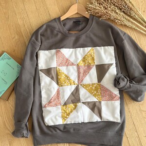 May include: A charcoal gray crewneck sweatshirt featuring a large, square, quilted star patch on the front. The patch is composed of white, mustard yellow, and floral-patterned fabric triangles arranged in a geometric star pattern with raw edges.