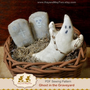 May include: A Halloween-themed basket contains a white ghost with blue eyes and a safety pin necklace, along with two gray tombstone-shaped pillows. One pillow has a cross, and the other has a skull and crossbones. The basket is filled with dried moss.