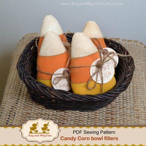 May include: A woven basket filled with four candy corn-shaped decorations. Each candy corn is made of white, orange, and yellow fabric and tied with twine. Small round tags with the words "Candy Corn" are attached to each piece. The basket is sitting on a woven surface.