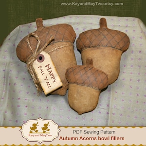 May include: Three fabric acorn bowl fillers in shades of brown and tan. One acorn has a tag that reads "Happy Fall Y'all". The acorns have a textured appearance and are arranged on a patterned surface. The image also includes the text "Autumn Acorns bowl fillers".