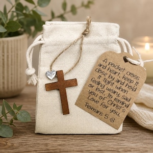 May include: A small off-white fabric drawstring pouch featuring a brown wooden cross charm and a silver heart charm labelled love. A brown paper tag is attached, printed with the text: A pocket cross and heart, keep it close by and hold it tight when feeling weary, sad or lost, to remind you of Christ's Love for you. Romans 5:8.