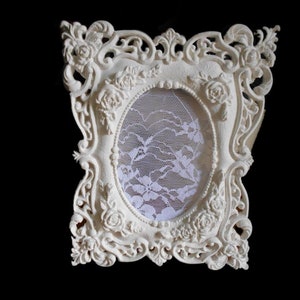 May include: A white ornate picture frame with a lace insert. The frame has a detailed design with floral accents.