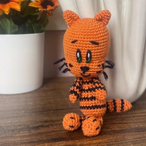 May include: A handmade orange and black crochet tiger figurine with green eyes and black whiskers. The tiger has black stripes and is sitting on a wooden surface. A white vase with orange flowers and a pot of sunflowers are in the background.