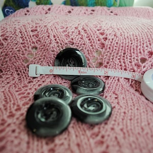 May include: A collection of black and gray swirl-patterned buttons on a pink knitted fabric. A white measuring tape shows 2 inches. The buttons are round and glossy. The knitted fabric has a delicate, openwork design.