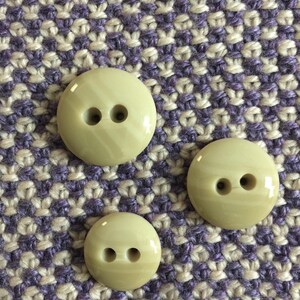 May include: Three beige buttons with two holes each. The buttons are on a purple and beige patterned fabric.