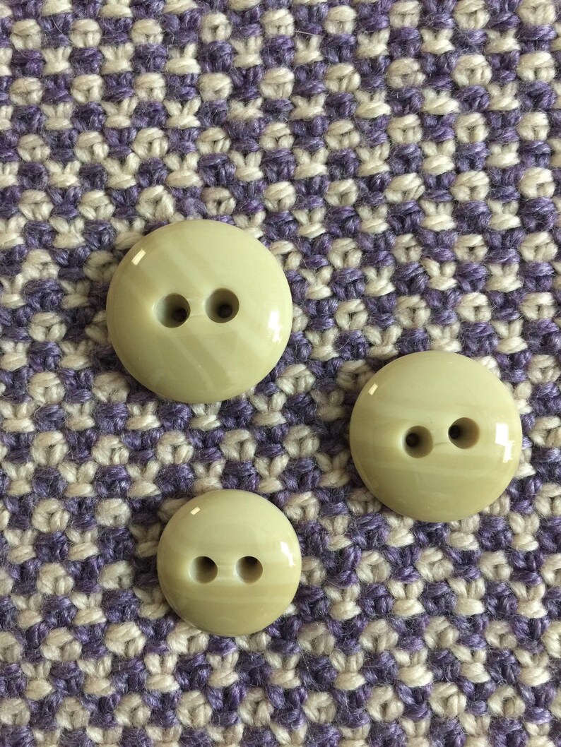 May include: Three beige buttons with two holes each. The buttons are on a purple and beige patterned fabric.