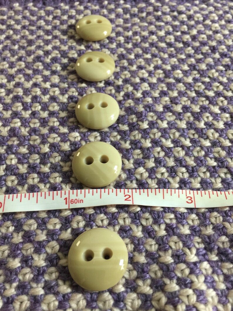 May include: Five beige plastic buttons with two holes each, lined up vertically on a purple and beige patterned fabric. A white tape measure with red markings is placed below the buttons.