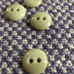 May include: Five beige plastic buttons with two holes each. The buttons are round and have a slight shine. They are arranged in a row on a purple and white patterned fabric.