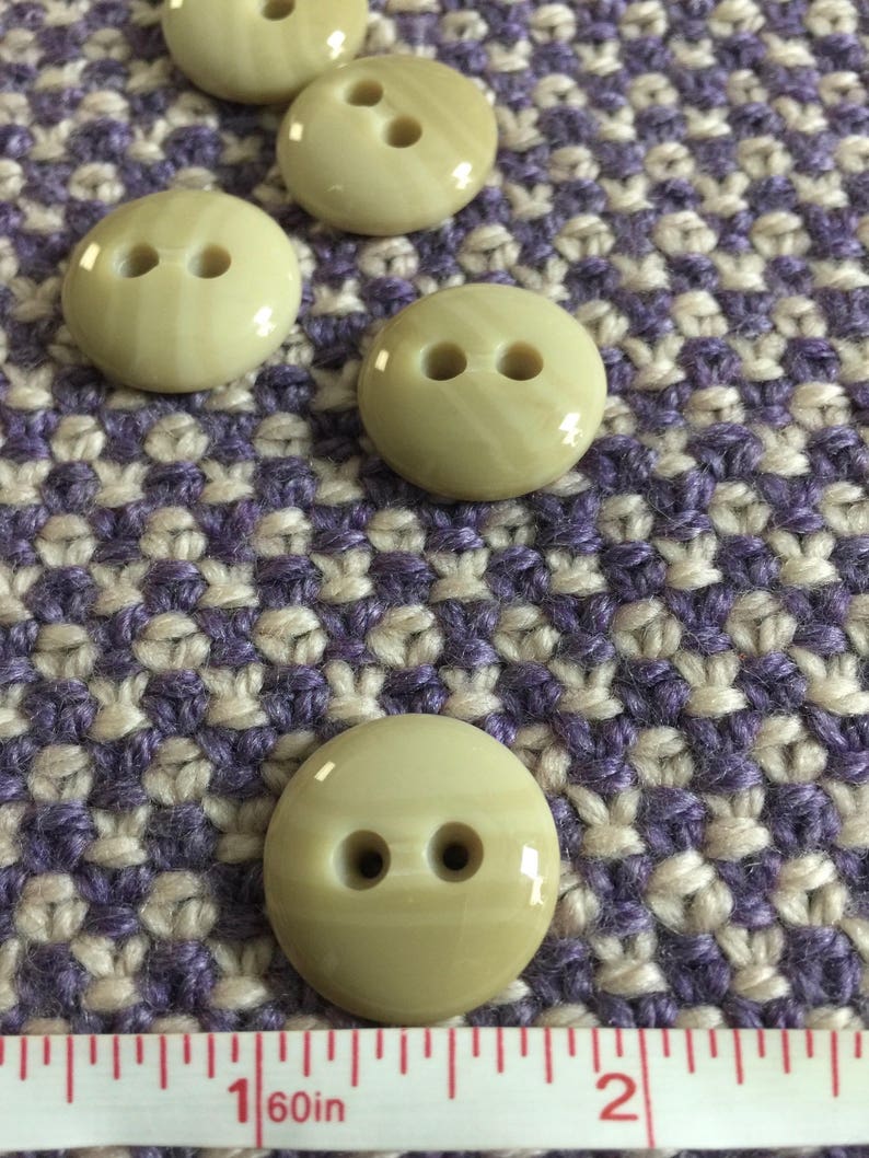 May include: Five beige plastic buttons with two holes each. The buttons are round and have a slight shine. They are arranged in a row on a purple and white patterned fabric.