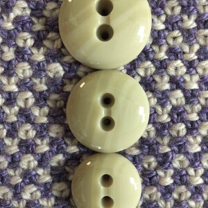 May include: Three beige plastic buttons with two holes each. The buttons are arranged in a vertical stack, with the largest button at the top and the smallest button at the bottom.