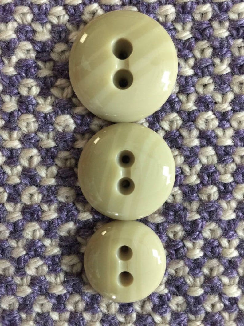 May include: Three beige plastic buttons with two holes each. The buttons are arranged in a vertical stack, with the largest button at the top and the smallest button at the bottom.