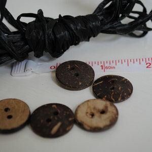 May include: Five brown coconut shell buttons with two holes each. The buttons are arranged in a semi-circle on a white surface. A black string and a white measuring tape are visible in the background.
