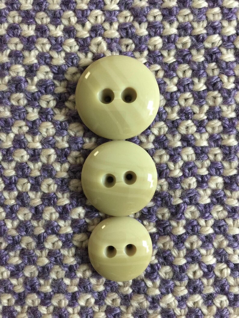 May include: Three round, beige buttons with two holes each. The buttons are arranged vertically, with the largest button at the top and the smallest button at the bottom.