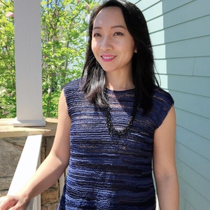 May include: A woman with long black hair is wearing a blue and black striped knit top with a black necklace. She is standing in front of a light blue wall.