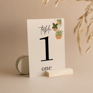 Puede incluir: Una tarjeta blanca con el texto "table 1 one" impreso en ella. La tarjeta está decorada con un dibujo de tres plantas en macetas colgantes. La tarjeta está apoyada en un pequeño bloque de madera.