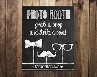 May include: Black chalkboard sign with white text that reads "PHOTO BOOTH grab a prop and strike a pose!" with a bowtie, mustache, and glasses drawn below. The hashtag #MrAndMrsJones is at the bottom of the sign.