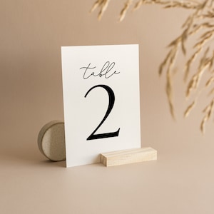 May include: A white table number card with the word "table" in a script font and the number "2" in a bold, black font. The card is displayed on a small wooden stand. A small, round, textured object is to the left of the card.