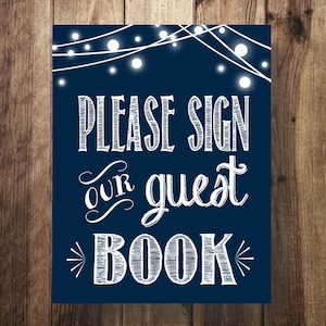 May include: Navy blue sign with white text that reads "PLEASE SIGN our guest BOOK". The sign has a string of white lights at the top. The background is a wooden texture.