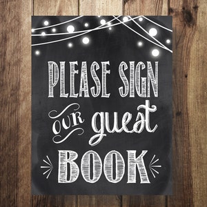 May include: Black chalkboard sign with white text that reads "Please sign our guest book". The sign is decorated with white string lights.