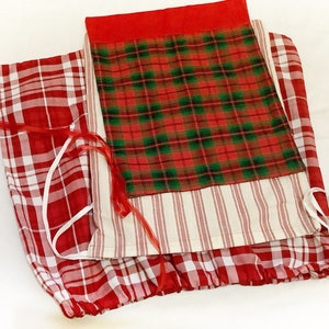 May include: A folded textile item featuring a patchwork design. The item incorporates red and white plaid, a red and green plaid, and a cream fabric with red stripes. Red ribbons and white ties are visible. The item appears to be a decorative home textile.