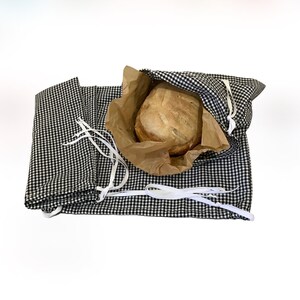 May include: A loaf of bread in a brown paper bag inside a black and white checkered bread bag. The bag has white drawstrings and is open, revealing the bread. The bread bag is laying on a white surface.