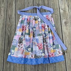 May include: A colorful patchwork apron with a blue waistband and ties. The apron features a variety of floral, striped, and patterned fabric squares in shades of pink, blue, yellow, and green. The apron is designed for kitchen use.