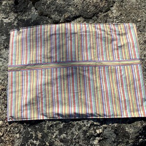 May include: A rectangular fabric placemat featuring a vibrant vertical striped pattern in shades of yellow, red, blue, green, and white. The textile includes a distinct horizontal band across the centre, also decorated with matching thin stripes.