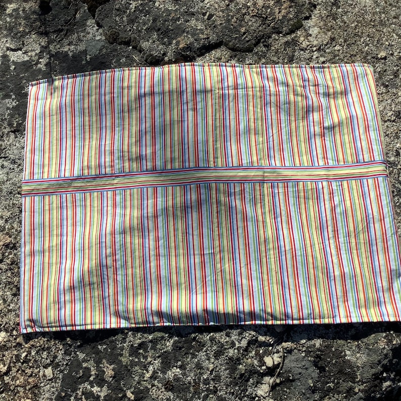 May include: A rectangular fabric placemat featuring a vibrant vertical striped pattern in shades of yellow, red, blue, green, and white. The textile includes a distinct horizontal band across the centre, also decorated with matching thin stripes.